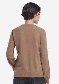 Frau mit kurzem dunklem Haar, die einen schlichten hellbraunen Pullover mit langen Ärmeln und dunklen Hosen trägt, von hinten vor einem weißen Hintergrund.