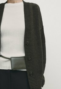 Tummanvihreä neulottu neuletakki, jossa resorit reunuksissa ja napillinen kiinnitys, päällä valkoinen resorinen paita. Pieni tumma vyölaukku vyötäröllä.