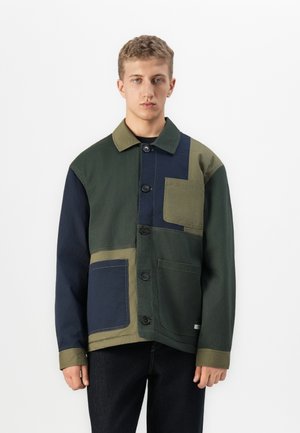 Young man wearing a color-blocked jacket with green, navy, and olive patches, featuring button closures and three front pockets.