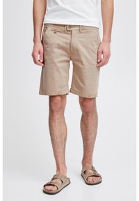 Homme portant des shorts beige jusqu'aux genoux et des sandales beiges, se tenant contre un fond clair uni.