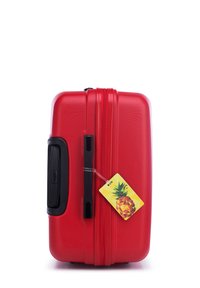 Valise rigide rouge avec une finition texturée, zip et poignée noirs. Une étiquette en forme d'ananas jaune est attachée.