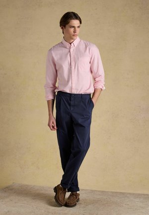 Young man wearing light pink striped button-up shirt, navy trousers, and brown shoes poses with one hand in pocket against beige backdrop.