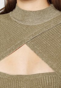 Gros plan sur une femme portant un haut en tricot côtelé beige avec un col montant et une découpe triangulaire au niveau de la poitrine.