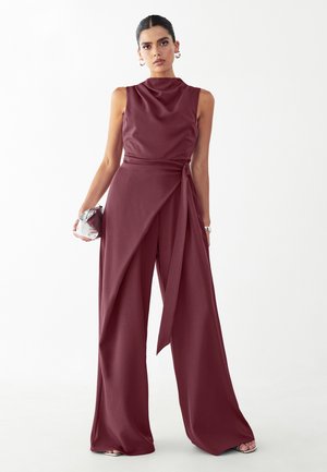 Kobieta ubrana w bezrękawnikowy kombinezon w kolorze bordowym z szerokimi nogawkami i drapowanym paskiem w talii, trzymająca małą metaliczną kopertówkę na białym tle.