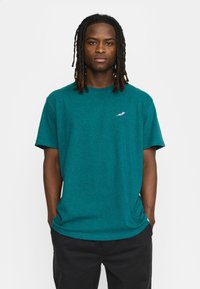 Teal katoenen t-shirt met een ronde hals en korte mouwen; voorzien van een klein, kleurrijk logo op de linkerborst. Gecombineerd met zwarte broeken.