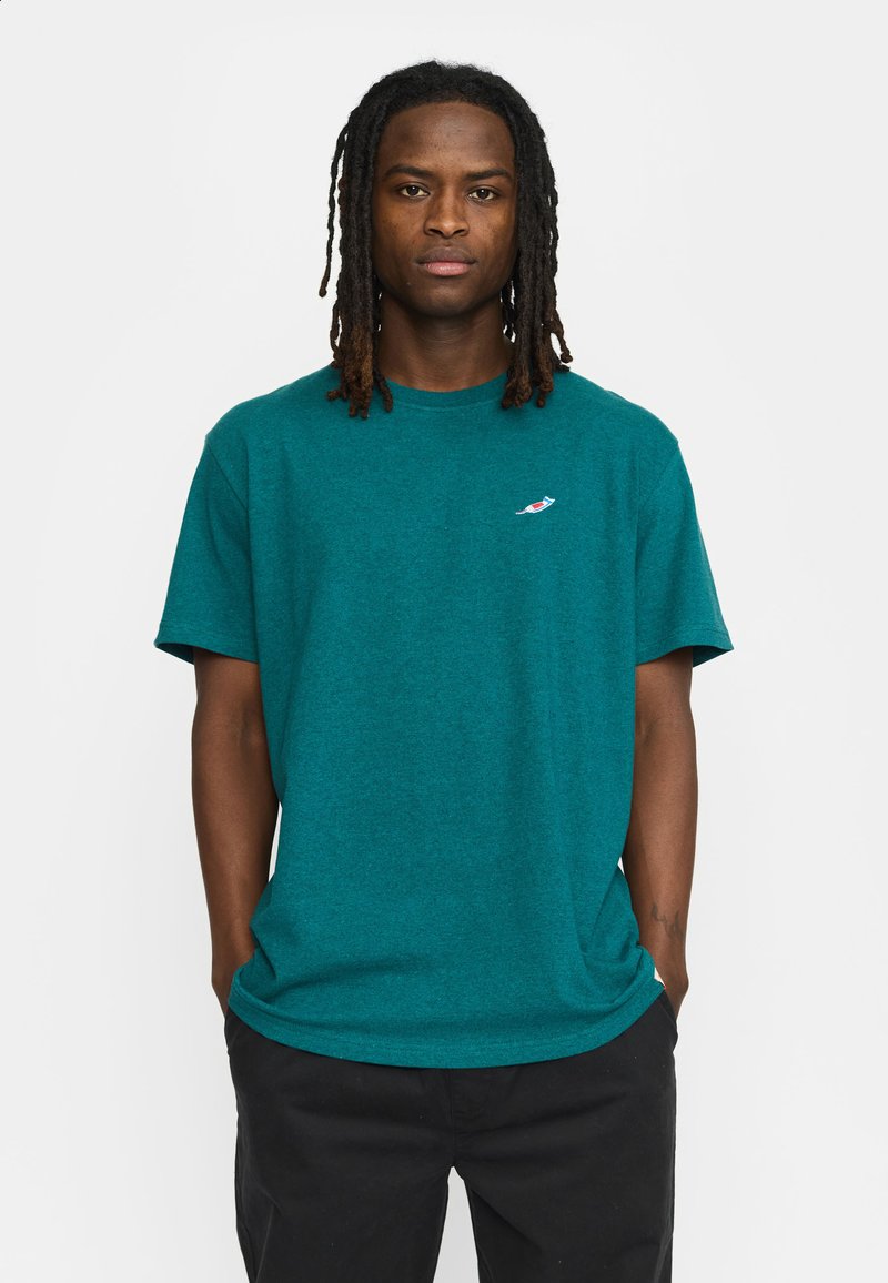 Teal katoenen t-shirt met een ronde hals en korte mouwen; voorzien van een klein, kleurrijk logo op de linkerborst. Gecombineerd met zwarte broeken.