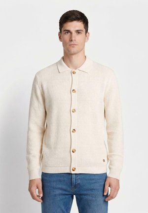 Mężczyzna w beżowym dzianinowym kardiganie z drewnianymi guzikami i niebieskich dżinsach, stojący na jednolitym białym tle.