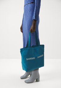 Τσάντα καμβά σε τόνους τιρκουάζ με το "SAM SøE" τυπωμένο σε λευκό, διαθέτοντας διπλούς ιμάντες. Φορεμένη παράλληλα με ένα μπλε ριγέ φόρεμα και γκρι μποτάκια στον αστράγαλο.