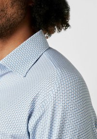 Hellblauses Hemd mit einem geometrischen Muster in dunklerem Blau. Der Stoff wirkt glatt mit einem leichten Glanz.