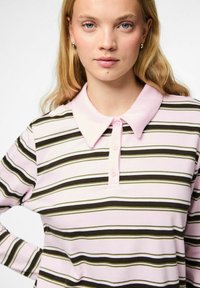 Šviesiai rožinė trumpomis rankovėmis polo marškiniai su horizontaliomis žaliomis ir juodomis juostelėmis. Yra smailus apykaklė ir trys mygtukai.