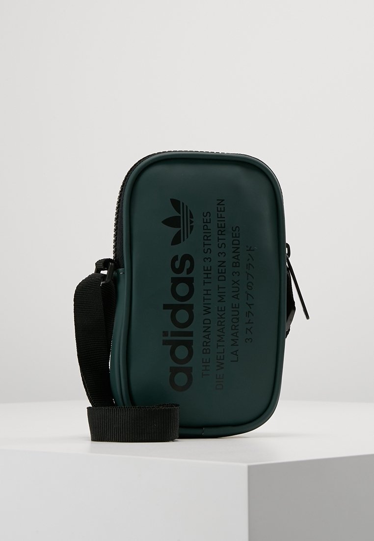 Zelená plastová taška přes rameno se zipovým uzávěrem, vyznačující se embosovaným černým textem a logem. Obsahuje černý popruh pro nošení.