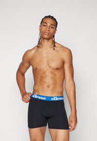 Svarta boxertrosor med en blå elastisk midja som har "ellesse"-logotypen. Designen är åtsittande, med en slät textur och en figurnära form.