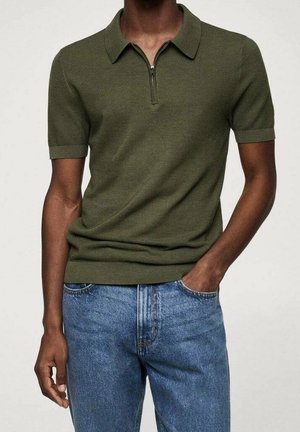 Personne portant un polo en maille vert olive à manches courtes avec une fermeture éclair sur le devant et un jean en denim bleu, une main dans la poche.