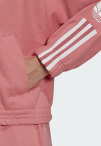 Sweatshirt rose avec des rayures blanches sur les poignets. Il présente une poche kangourou et des détails côtelés au bas et aux poignets pour une texture supplémentaire.