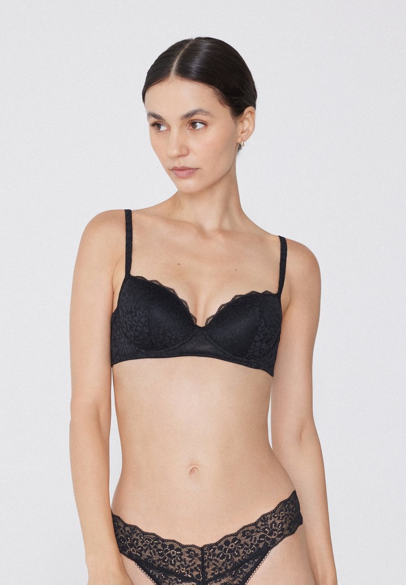 Sosten negro de encaje con bordes en forma de ondas, con un diseño acolchado y tirantes ajustables. Combinado con braguitas de encaje a juego.