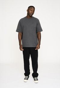 Grå T-shirt med korte ærmer og afslappet pasform, der har rund halsudskæring. Båret med sorte bukser og sorte sneakers med hvide snørebånd.
