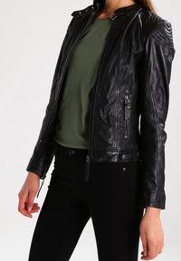 Zwarte leren jas met een slim fit ontwerp, ritssluitingen aan de voorkant, geribbelde schouders en zijzakken. Gecombineerd met een groene shirt en zwarte broek.