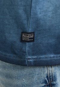 Camiseta azul de cuello redondo hecha de algodón suave. Presenta un dobladillo cosido y una etiqueta de tela que detalla "Petrol Ind" cerca de la costura inferior.