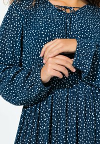 Gaun biru navy dengan polka dot putih, dilengkapi dengan leher yang berkumpul dan lengan lonceng panjang. Kain mengalir dengan detail lipitan di pinggang.