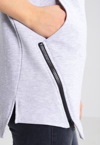 Light gray sweatshirt with a side zipper pocket, featuring a textured fabric and side slits at the hem, paired with dark jeans.