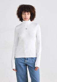 Witte lange mouw turtleneck top in ribbelstructuur, met een kleine logoprint op de borst; gecombineerd met losse, blauwe jeans.