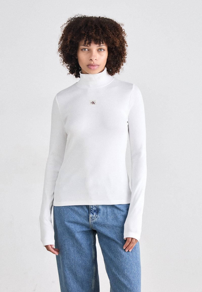 Witte lange mouw turtleneck top in ribbelstructuur, met een kleine logoprint op de borst; gecombineerd met losse, blauwe jeans.