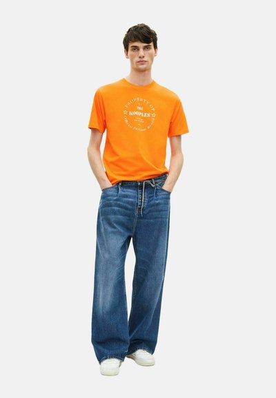 Oranje T-shirt met witte grafische print, gecombineerd met losse blauwe jeans en witte sneakers, welke een casual stedelijke stijl tentoonstelt.