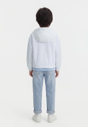 Kind trägt eine weiße Kapuzenjacke, hellblaue Jeans mit hochgekrempelten Säumen, weiße Socken und weiße Turnschuhe, steht mit dem Rücken zur Kamera.