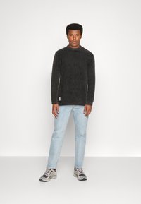 Dunkelgrauer, gerippter Pullover mit hohem Kragen, kombiniert mit hellblauen Jeans und mehrfarbigen Sneakern. Der Stoff wirkt weich und strukturiert.