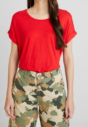 T-shirt rosso a maniche corte con orli arrotolati, abbinato a pantaloni cargo beige e verdi in camo con chiusura a bottone e tasche frontali.