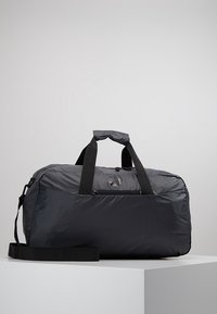 Musta pehmeä duffel-laukku, jossa on kaksi kantokahvaa, etupuolella vetoketjullinen tasku ja säädettävä olkahihna valkoisella alustalla.