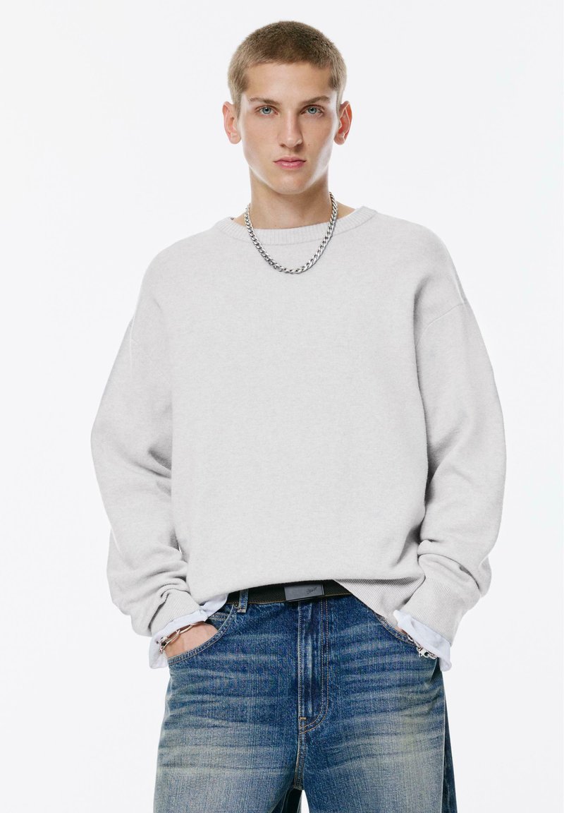 Hanorac oversized gri deschis, cu maneci lungi si decolteu rotund; asociat cu blugi din denim albastru, cu o textura usor deteriorata.