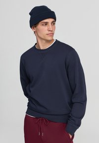 Felpa blu navy con polsini e orlo a coste, colletto a girocollo, indossata sopra una camicia bianca. Abbinata a pantaloni bordeaux e un berretto abbinato.