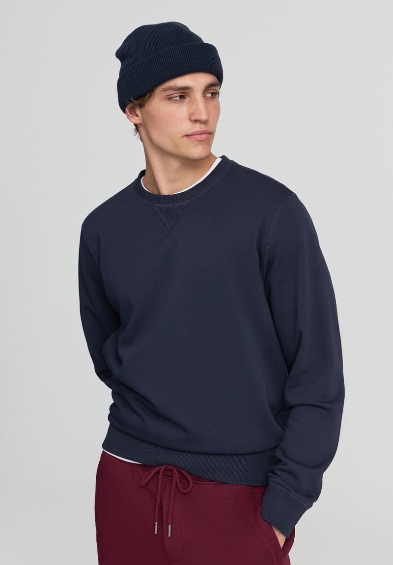 Felpa blu navy con polsini e orlo a coste, colletto a girocollo, indossata sopra una camicia bianca. Abbinata a pantaloni bordeaux e un berretto abbinato.