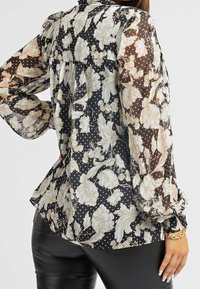 Blusa nera trasparente con motivi floreali e a pois, maniche lunghe a sbuffo e orlo svasato. Presenta un design posteriore leggermente arricciato.