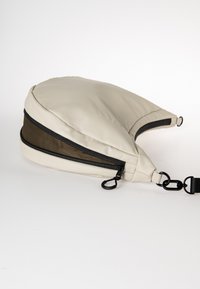 Cremefarbene Tasche mit einer weichen, gepolsterten Außenfläche. Verfügt über schwarze Reißverschlüsse, ein strukturiertes braunes Akzent und einen verstellbaren Gurt mit Hardware-Befestigungen.