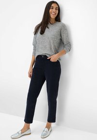 Femme appuyée contre un mur blanc, portant un pull gris à manches longues, un pantalon bleu foncé et des mocassins argentés, souriant avec une main dans la poche.