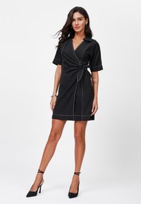 Black wrap dress with short sleeves, white contrast stitching, and a tie at the waist. Smooth texture and knee-length design.