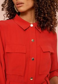 Femme avec des cheveux bouclés portant une chemise rouge à boutons avec deux poches avant et de grandes boucles d'oreilles en or, montrée de la menthe à la taille.