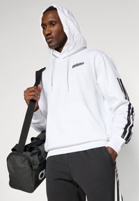 Sudadera blanca con logo negro y rayas negras en las mangas, combinada con pantalones gris oscuro. El modelo sostiene una bolsa de gimnasio negra con el logo.