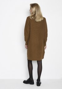 Robe pull en tricot marron avec des manches longues, fentes latérales et texture côtelée, associée à des chaussures noires épaisses et des collants.
