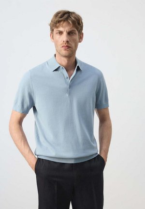 Hombre con cabello castaño claro que lleva una camisa tipo polo de manga corta azul claro y pantalones negros con las manos en los bolsillos, de pie frente a un fondo liso.