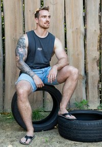 Black sleeveless shirt with logo, blue denim shorts, black sandals. Sitting on black tires, wooden background, sunlight on skin.