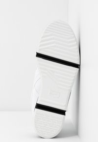 Semelle de chaussure blanche avec le logo Onitsuka Tiger, grip texturé et deux bandes de caoutchouc noires horizontales sur fond blanc.