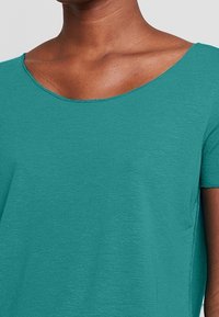 T-shirt teal a maniche corte con scollatura ampia, realizzato in un tessuto morbido e elasticizzato; cuciture visibili e texture liscia.