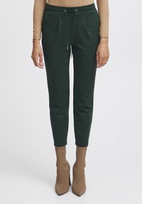 Vrouw in donkergroene tapered sweatpants met drawstring, tan lange mouwen top en tan puntige soklaarzen, staand tegen een witte achtergrond.