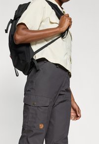 Svart ryggsäck med texturerat tyg, bärs på en beige skjorta och grå cargo-byxor med sidofickor och en logotypmärke.