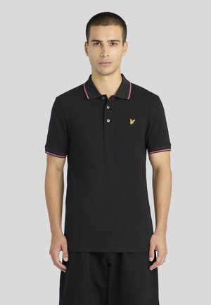 Homme portant un polo noir avec un liseré rose au col et aux manches, et un petit logo d’oiseau jaune sur la poitrine, debout devant un fond uni.