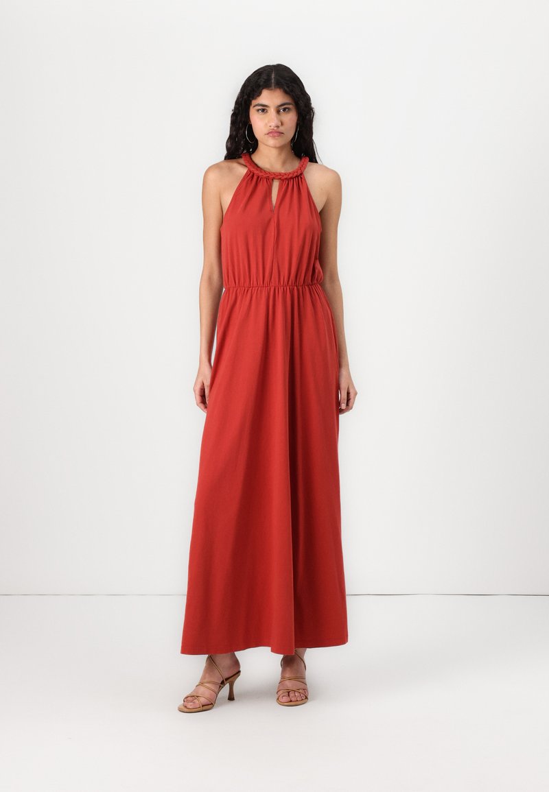 Vestido maxi em tecido vermelho-alaranjado com decote halter, detalhe em buraco e cintura elástica. Combinado com sandálias beiges de tiras. Textura suave.