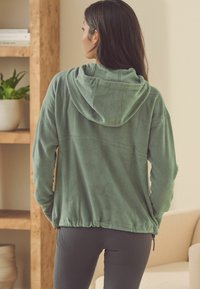 Grøn fleece hættetrøje med snoretræk i hætten, elastisk bund og lange ærmer. Blød tekstur, afslappet pasform med syninger på bagsiden.